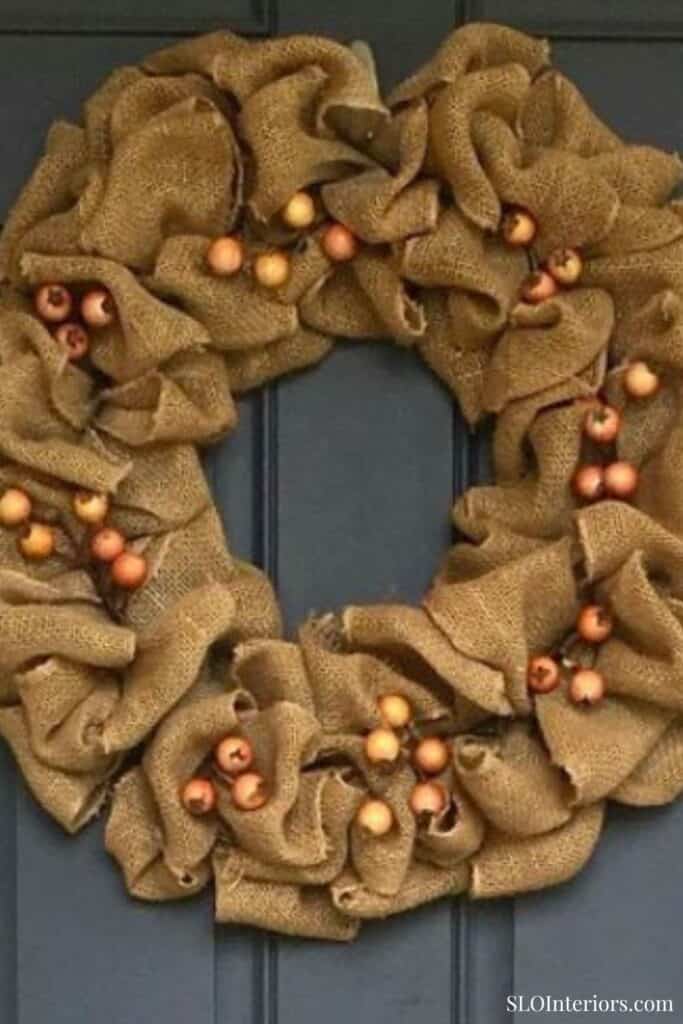 A festive burlap wreath decorated with copper and gold ornaments on a dark door.