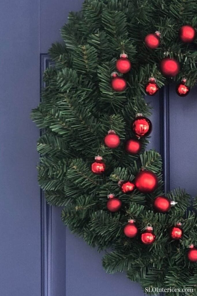A lush green Christmas wreath decorated with red ornaments hanging on a blue door.