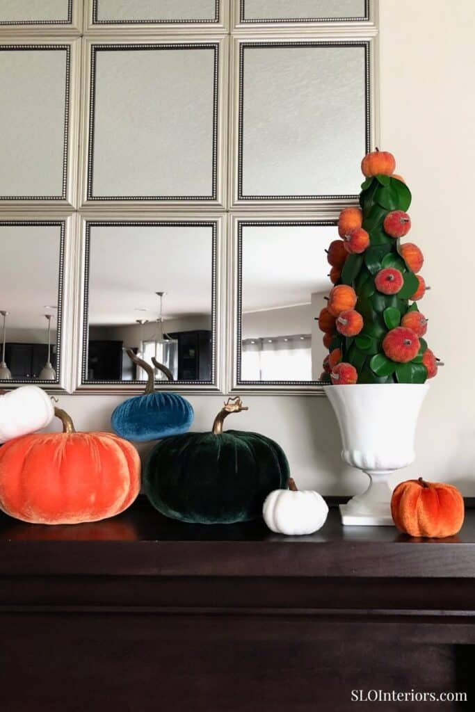 Pumpkin and apple decor on dark wooden table with mirror and wall panels in background.