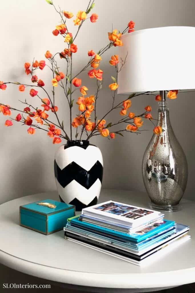 Vase with orange and yellow artificial flowers on a white side table with a silver lamp.