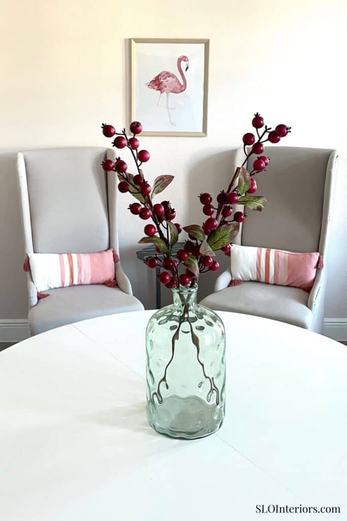 A lush red berry branch in a glass vase on a white table with two chairs and flamingo artwork.