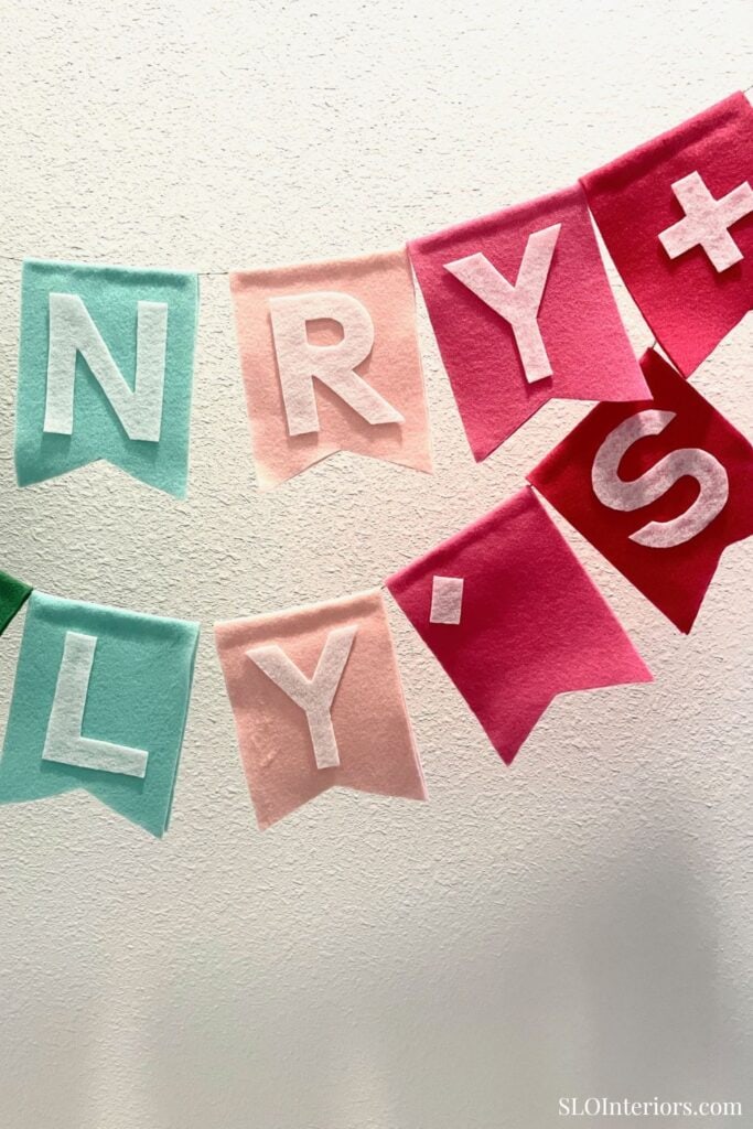 NRY+L Y+S festive felt banner with pastel and red colors on white wall.