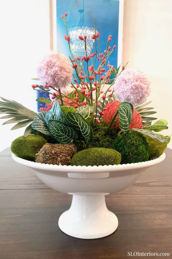 Tropical summer centerpiece in a white pedestal bowl with moss balls, pink flowers, coral stems, and green leaves.