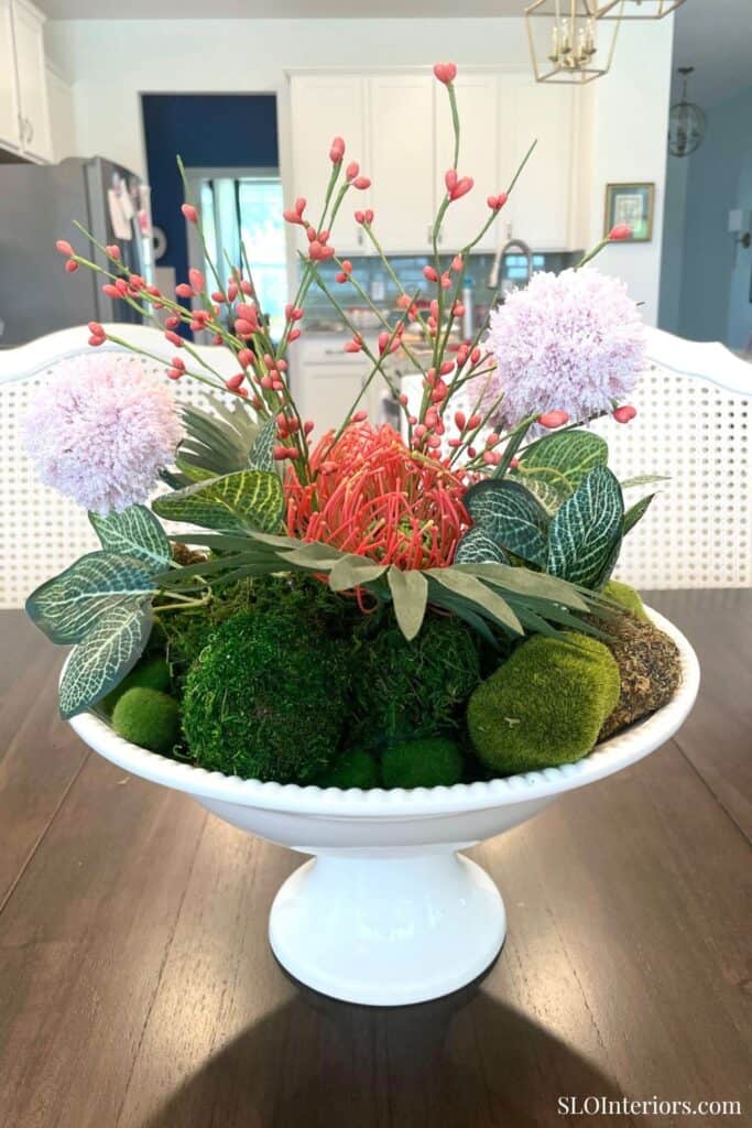 DIY summer table centerpiece with tropical greenery, pink blooms, moss spheres, and coral stems in a white footed bowl.