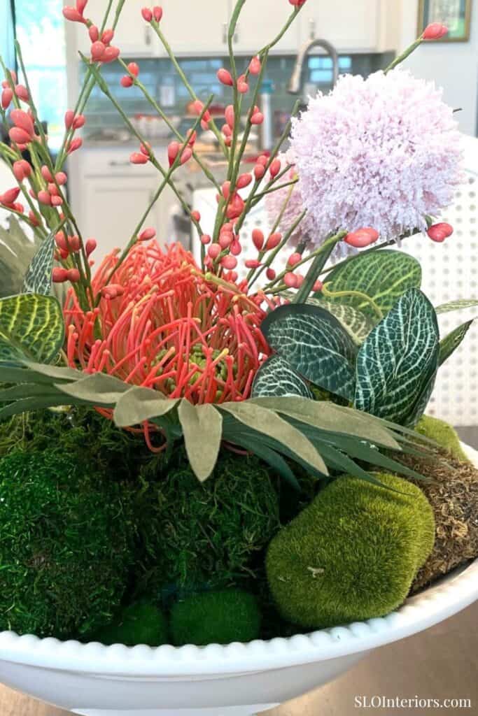 Colorful summer floral arrangement in a white compote bowl with moss, patterned leaves, pink flowers, and coral accents.