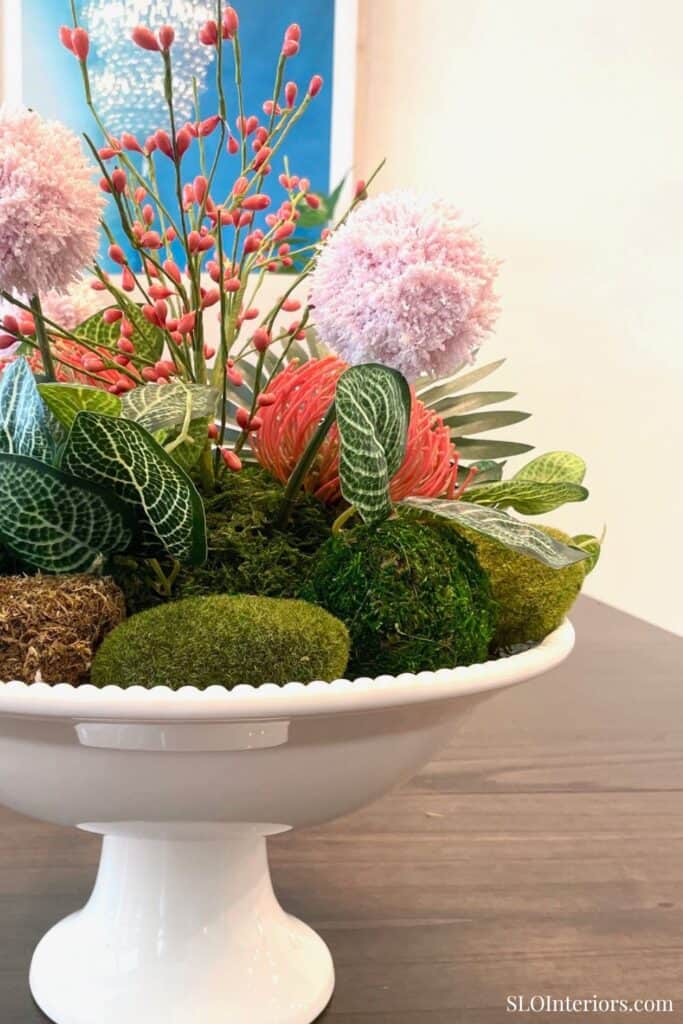 Tropical centerpiece for a summer table with moss balls, leafy greenery, pink pom-pom blooms, and coral branches.