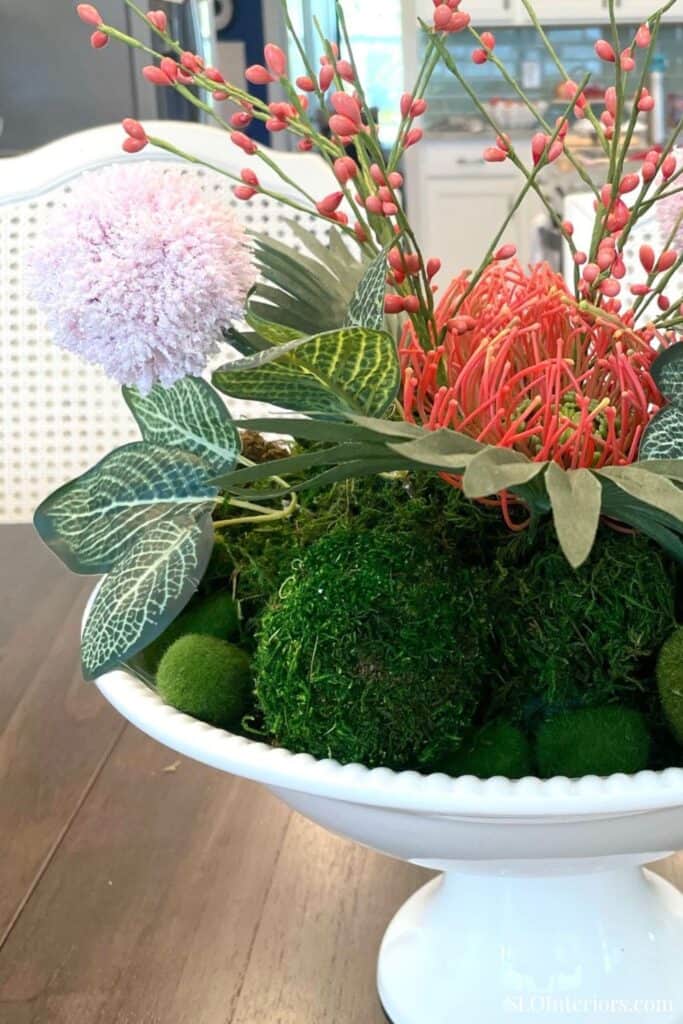 Summer table centerpiece in a white pedestal bowl featuring moss, tropical leaves, pink florals, and coral details.