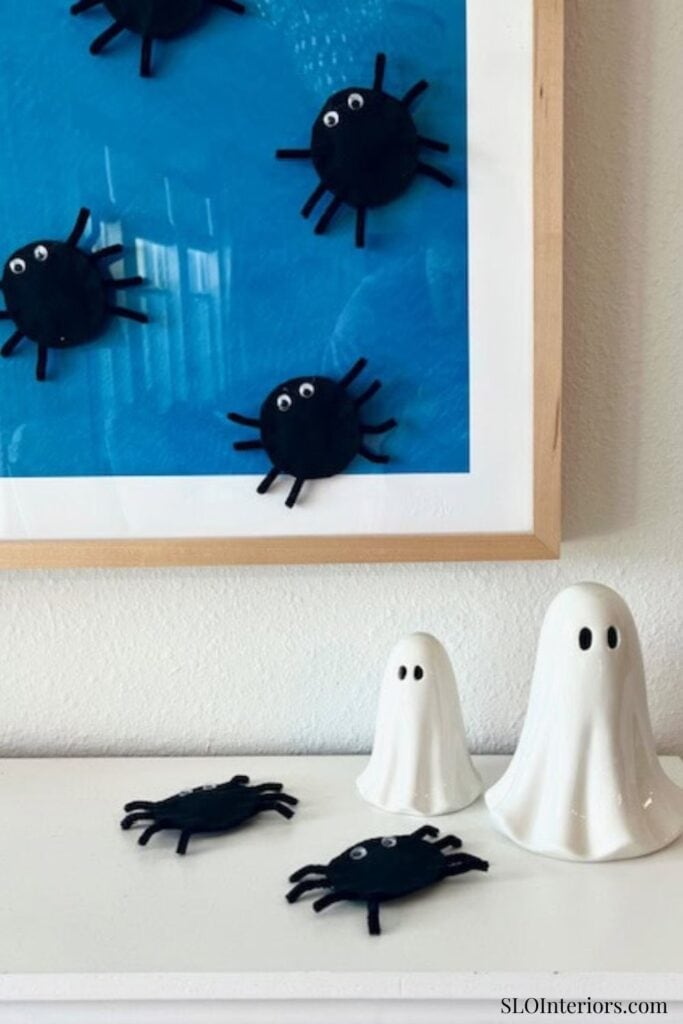 Cute black spider decorations on a blue framed wall art with ghost-shaped figurines on a white surfa.