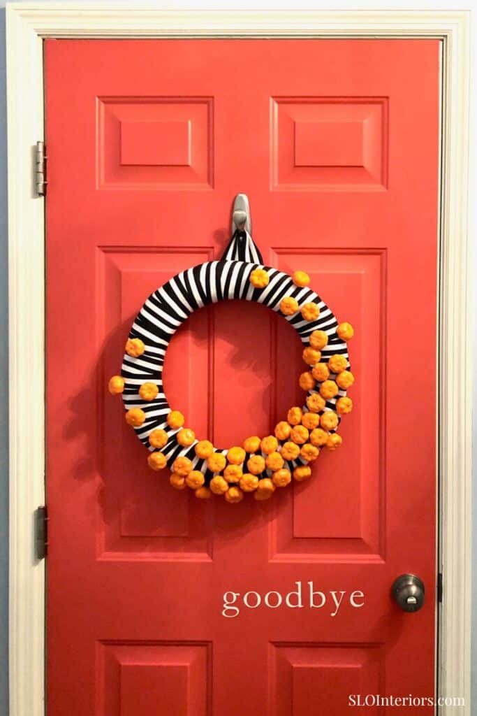 A colorful striped door wreath with orange pom-poms hanging on a coral door.