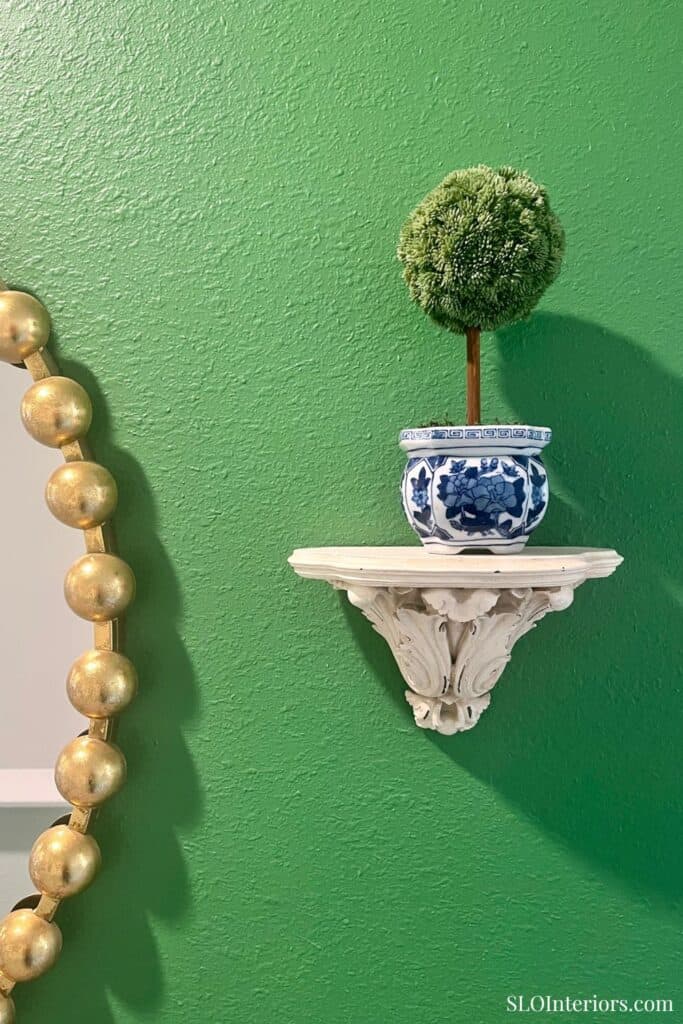 A lush green wall with a decorative white wall shelf holding a potted topiary plant.