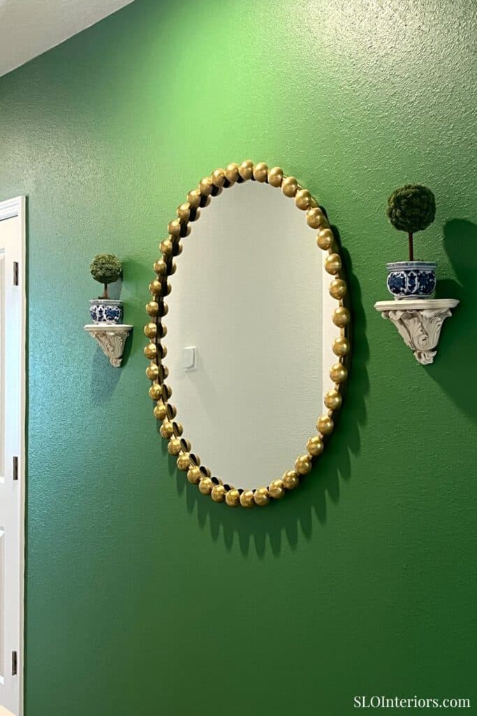 Elegant oval mirror with gold beaded frame on green wall, decorative wall shelves with potted plants.