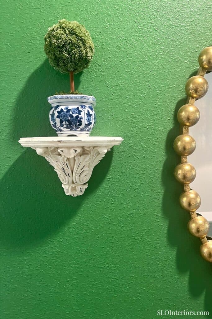 Decorative green wall with white ornate shelf, potted plant, and gold bead garland.