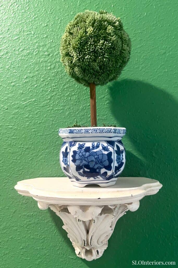 A lush green topiary in a decorative blue and white ceramic pot on a white ornate wall shelf.