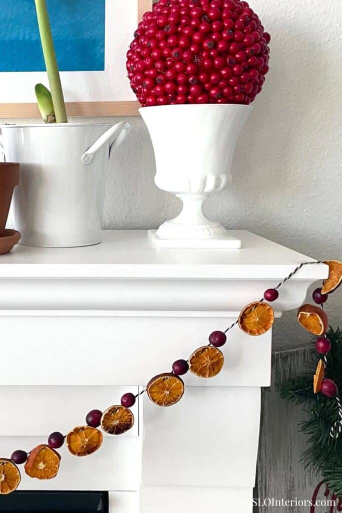 Red berry centerpiece in a white vase on a white mantel with holiday decor and a potted plant.