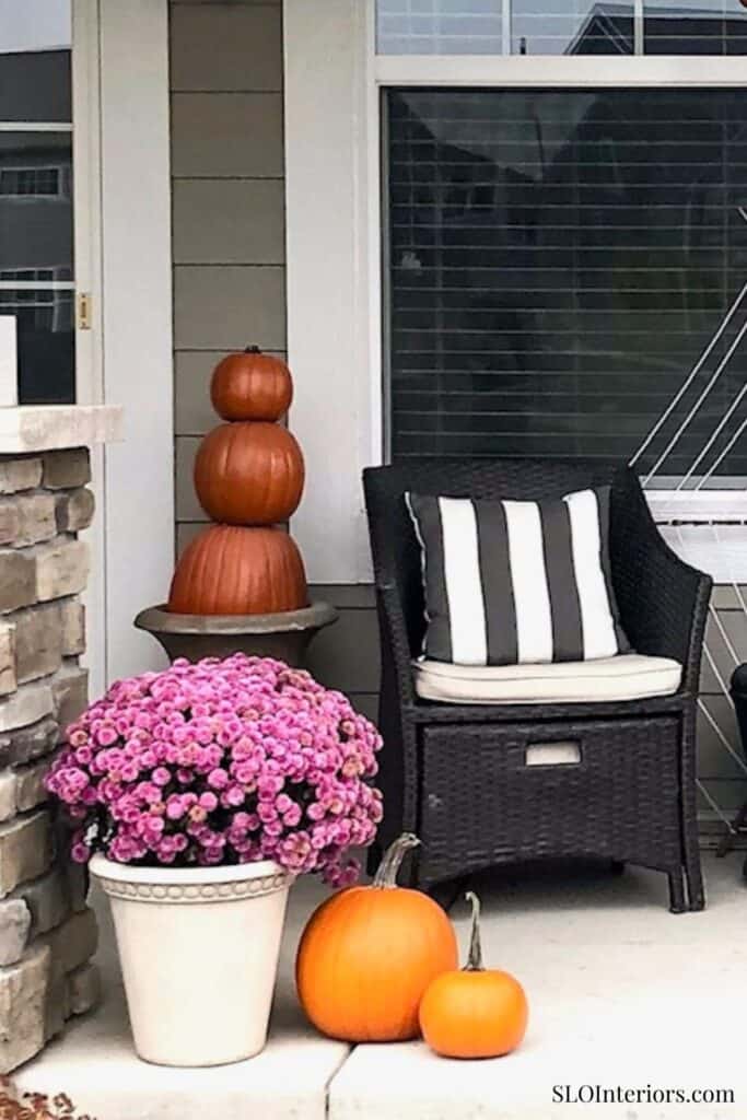 Pumpkin and pink chrysanthemum arrangement on porch with black wicker chair and fall decor.