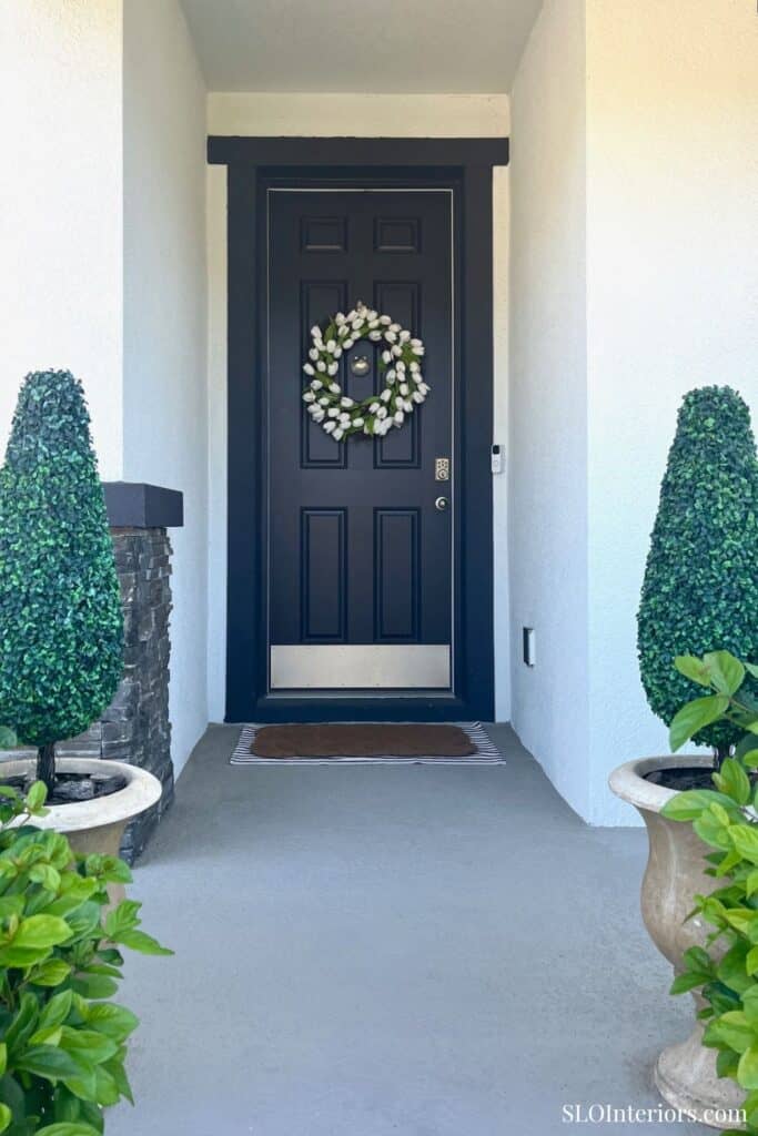 Elegant black front door with floral wreath, surrounded by lush greenery and potted plants.