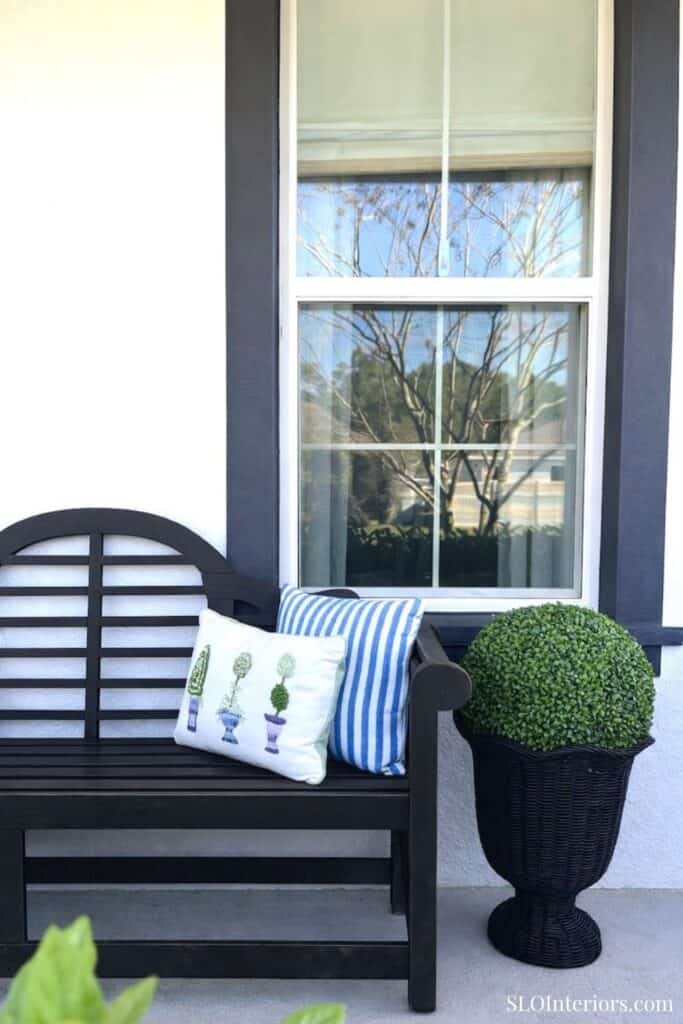 A lush potted plant and decorative pillows on a black outdoor bench by a window.