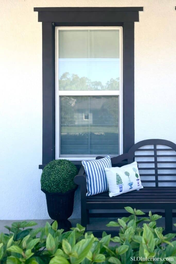 A lush outdoor seating area with a black bench, decorative pillows, and a window with black trim.