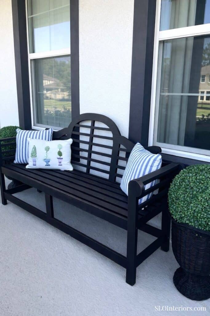 Black wooden outdoor bench with striped and decorative pillows on a porch with large windows.