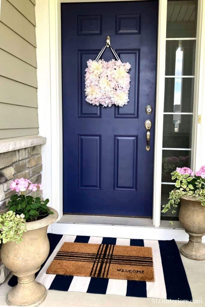 A lush floral wreath hanging on a navy front door with potted plants on each side.