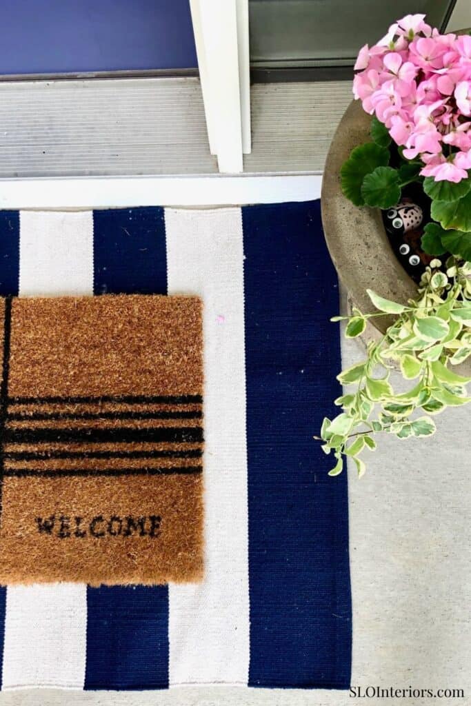Welcome doormat with navy and white stripes at entrance door, floral pot nearby.