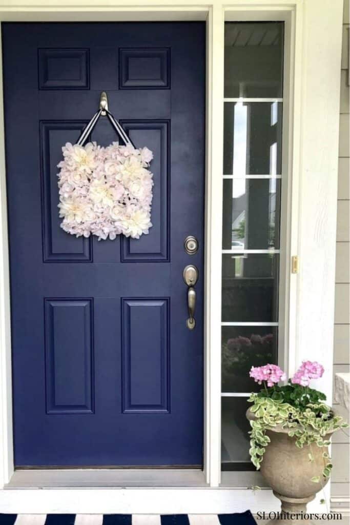 A colorful floral wreath hanging on a navy blue front door with a side window and potted pink flower.