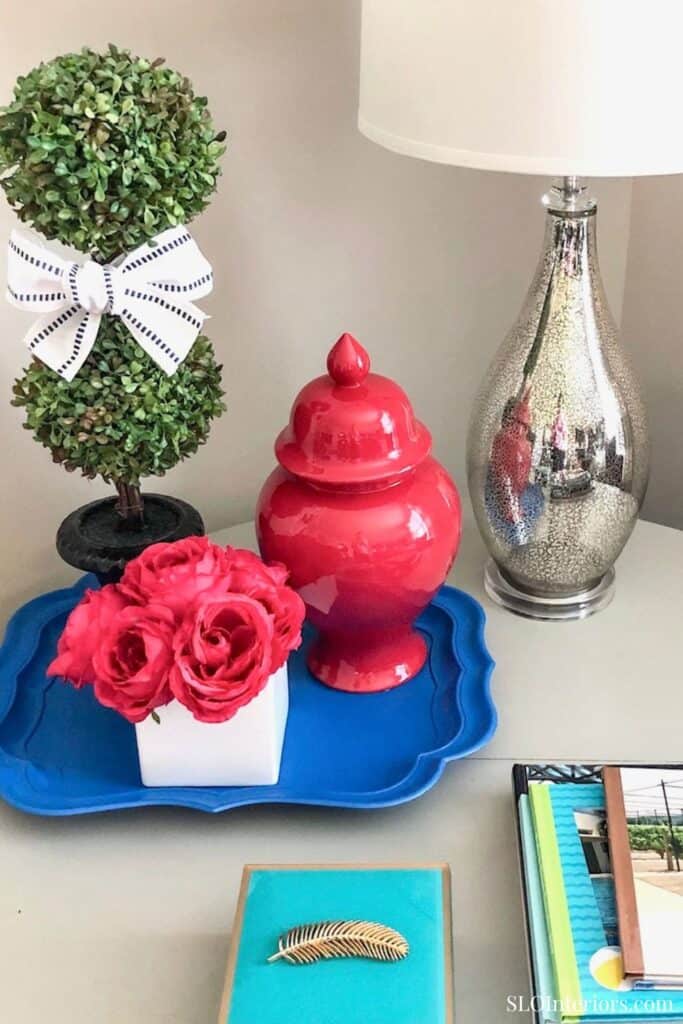 Artificial topiary plant with bow, red ceramic vase, silver lamp, and decorative items on a white ta.