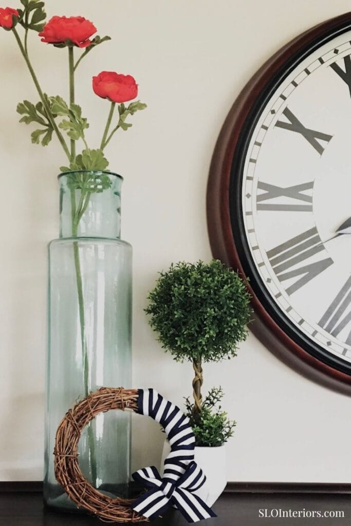 A lush green topiary plant with a striped bow in a white pot, next to a large wall clock and glass v.