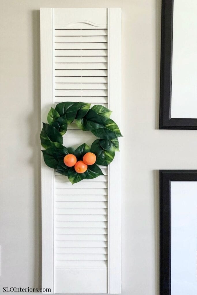 A lush green wreath with orange accents hanging on a white louvered door.