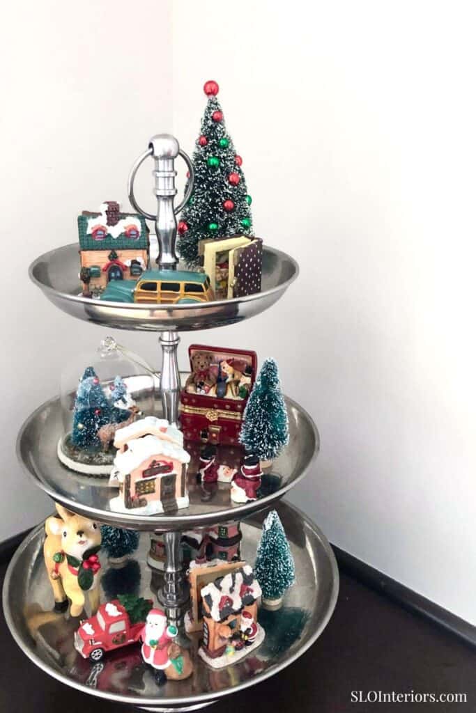 Festive holiday miniature village with Christmas trees and decorations on a tiered stand.