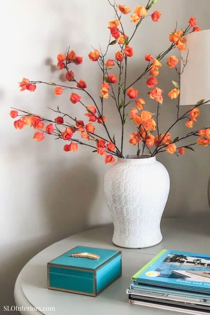 Elegant white vase with orange flowering branches on a modern side table, interior decor.
