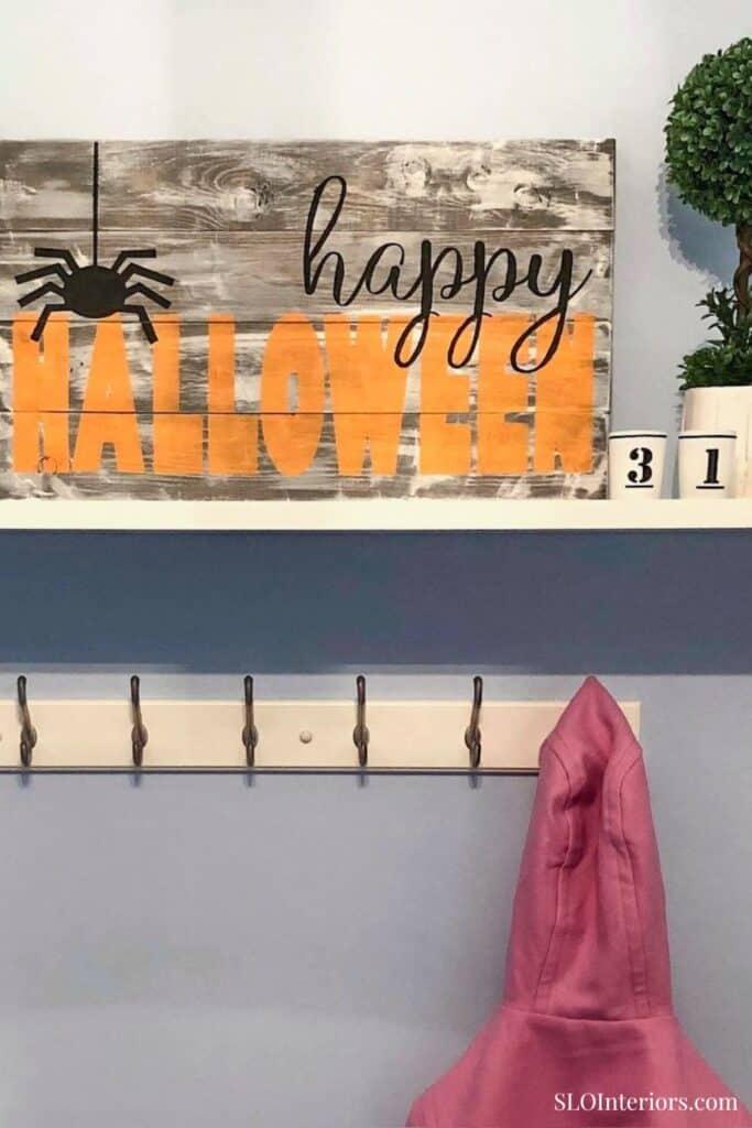 Happy Halloween wooden sign with spider decoration and pink coat hanging on wall hook.