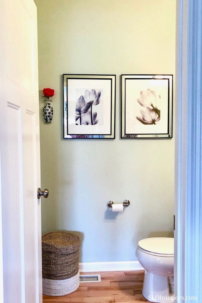 A lush bathroom with framed floral art, a decorative vase, and a woven laundry basket.