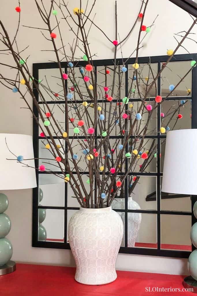White urn centerpiece with sticks and colorful pom poms on a red table