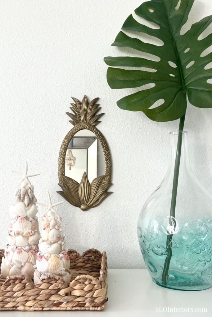 A lush Monstera leaf in a glass vase with decorative seashells and a pineapple-shaped mirror.