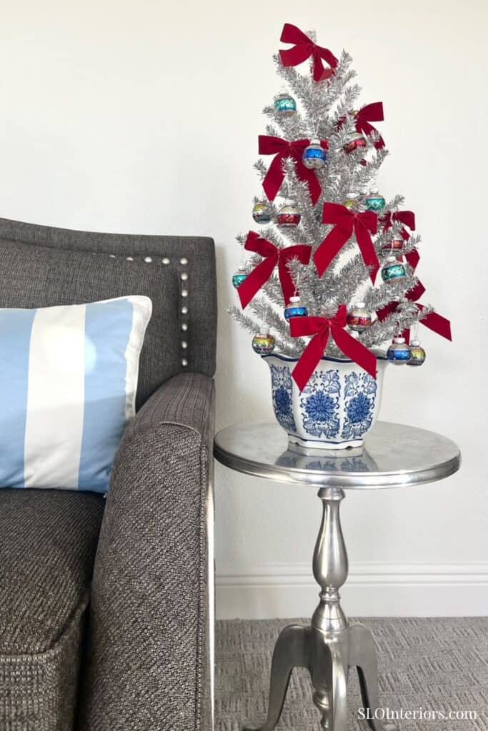 A Christmas tree decorated with red ribbons and blue ornaments next to a gray sofa and silver side t.