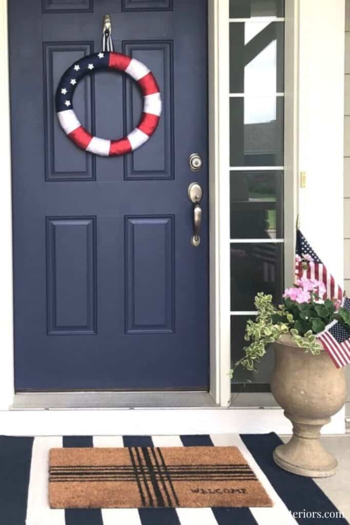 Patriotic wreath on navy front door with American flags and flower pot, welcoming entryway.