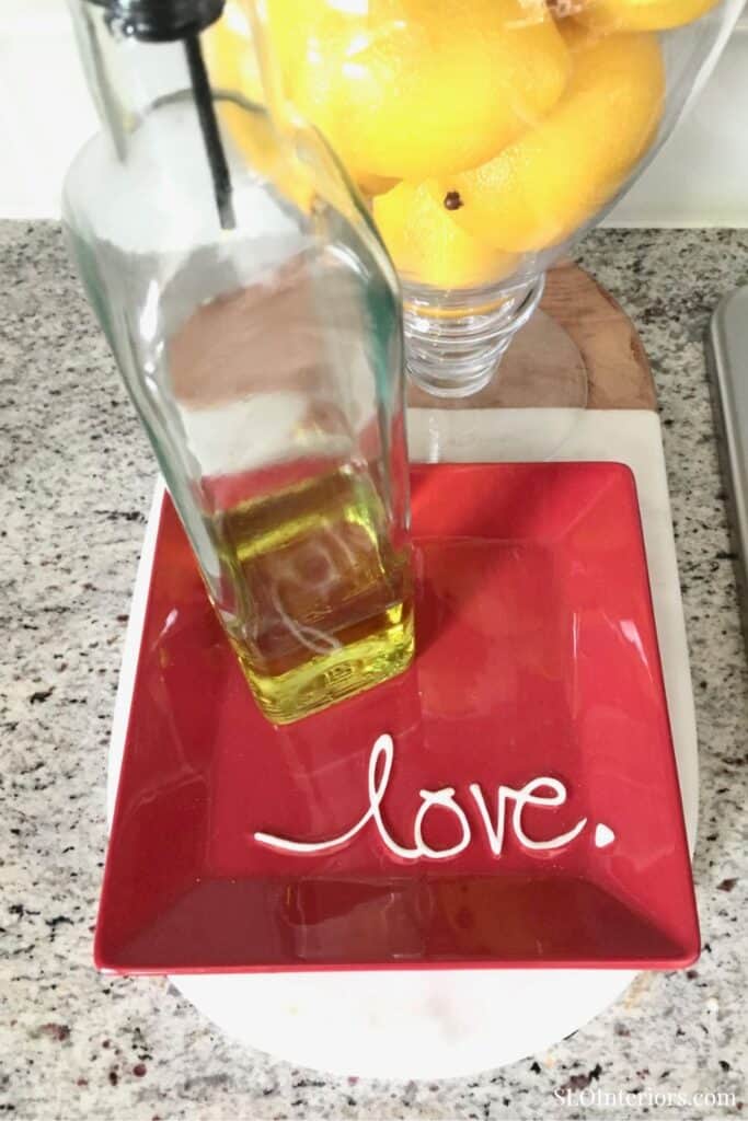 A sleek glass bottle of olive oil on a red "love." plate with lemons in a glass bowl.