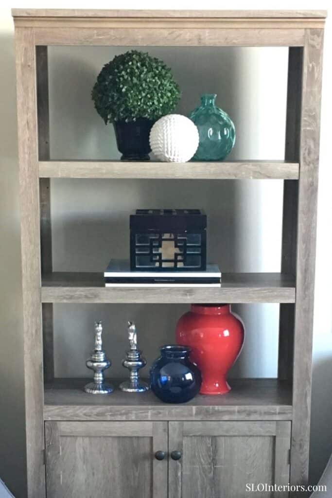 Artificial green topiary, decorative vases, and modern accents on wooden shelf.