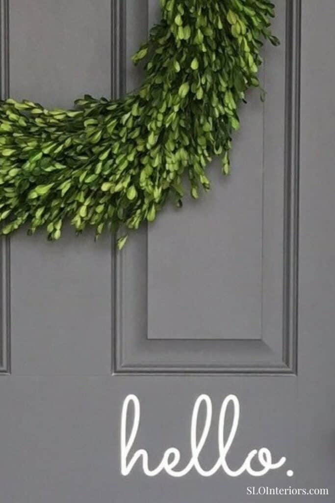 Green leafy wreath hanging on a gray door with 'hello' sign underneath.