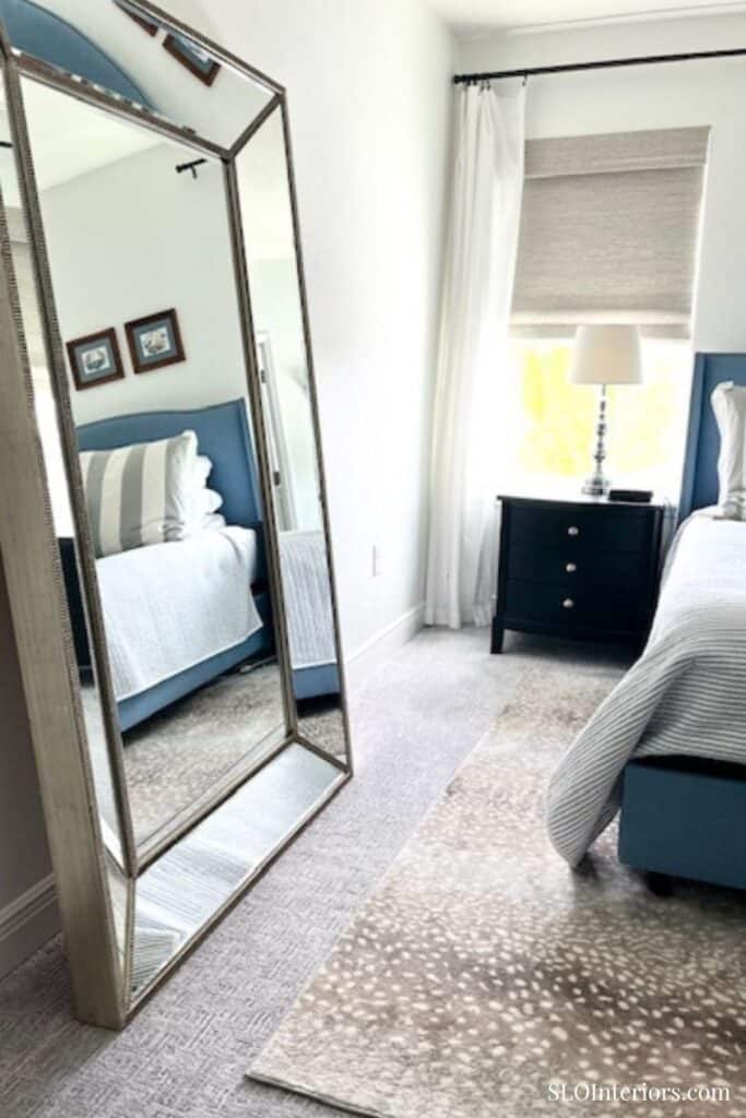 A modern bedroom with a large mirror, blue bed, and natural light from a window.