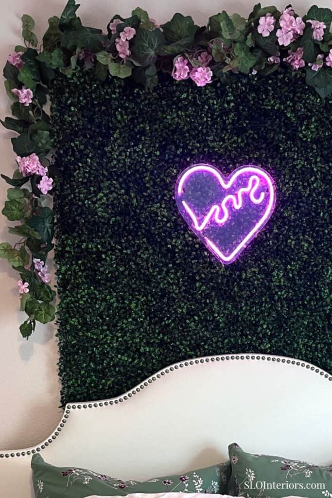 Decorative wall with pink flowers, green foliage, and neon love sign at SLO Interiors.