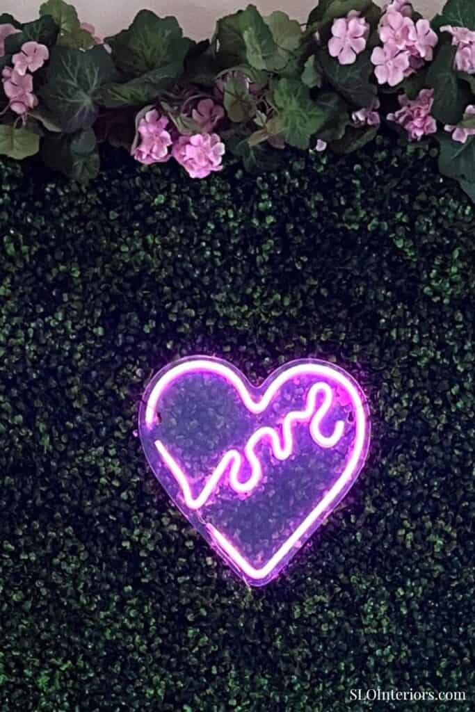 Pink neon heart sign on green hedge wall with pink flowers at top.