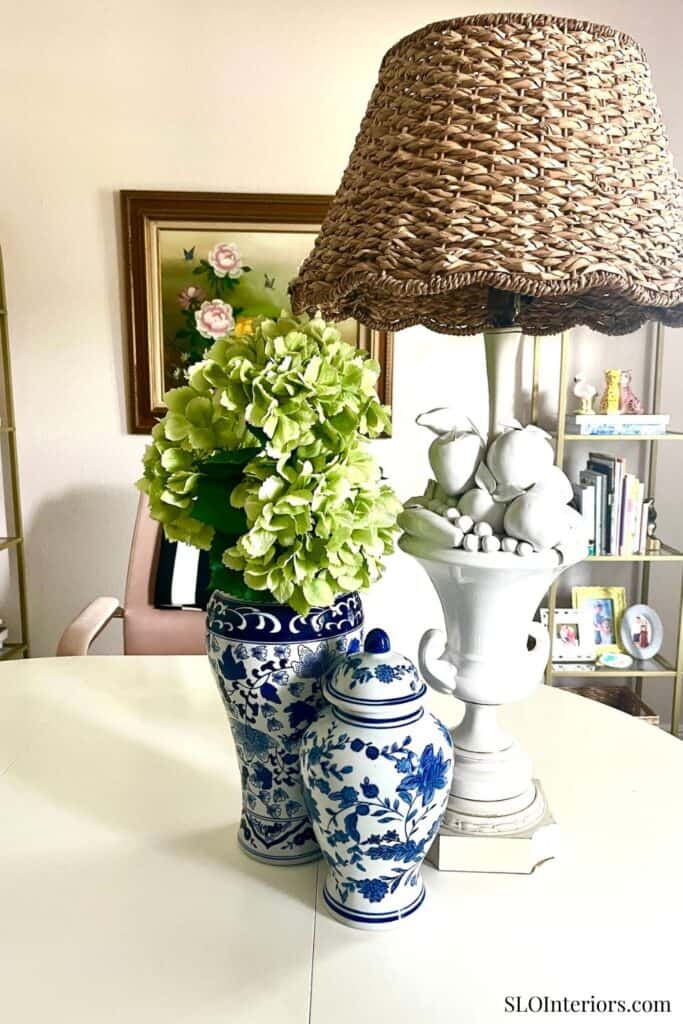 Decorative vases with green hydrangeas and a wicker lampshade on a white table.