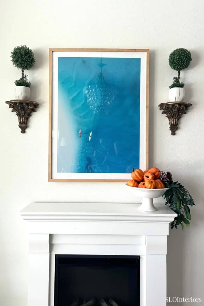 Decorative wall art above a white fireplace with plants and pumpkins for fall decor.