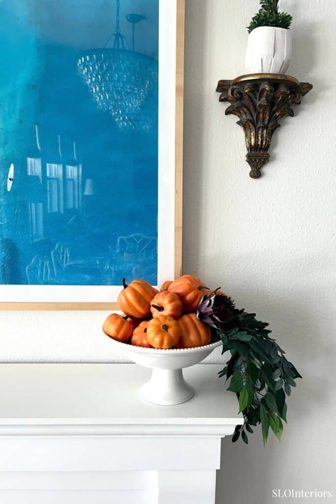 Decorative fall pumpkins and greenery on a white table with wall art and shelf.