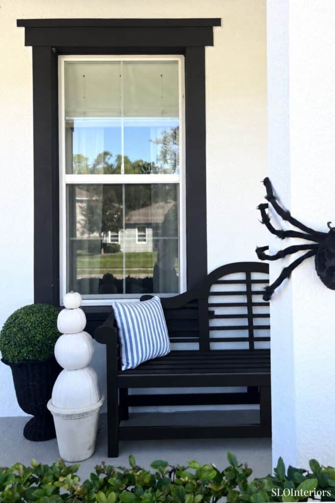 Black outdoor bench with striped pillow under window with black trim and spider decor.